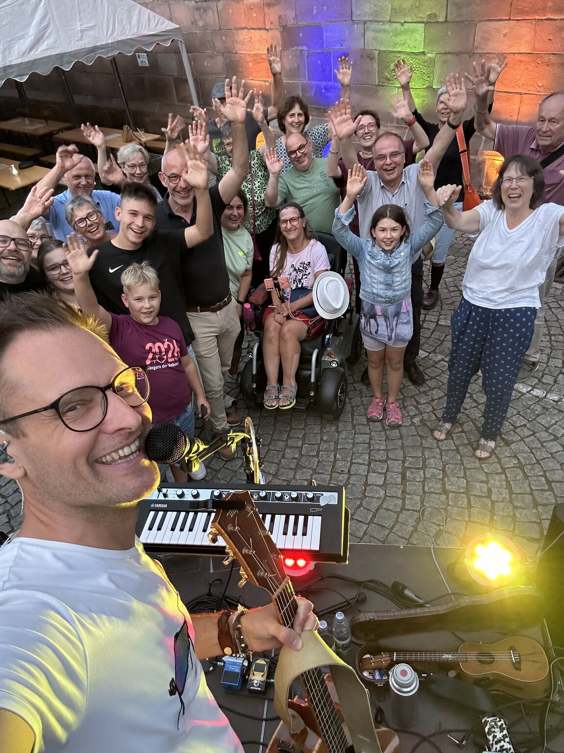 Selfie mit Sebastian Niklaus und den Zuhörenden beim Jakobsfest der St. Jakobskirche in Gernsbach. Auf dem Foto sind Gitarre, Ukulele, das Mikrofon und ein kleines Piano zu sehen. Die Bühne ist bunt beleuchtet. 